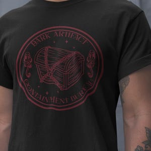 May include: Black t-shirt with a red circular graphic. The graphic features a treasure chest chained shut, surrounded by the words "DARK ARTIFACT CONTAINMENT BUREAU". Two stylized keys flank the chest.