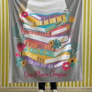 May include: Gray blanket featuring a colorful illustration of a stack of books with floral accents. The books are in various colors, including yellow, blue, and purple. The text "One More Chapter" is written in pink.