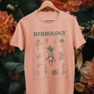 May include: A heather peach short-sleeve t-shirt featuring the word HERBOLOGY in dark green text above a square graphic containing various botanical illustrations and a central mandrake root figure printed in a matching dark green ink.