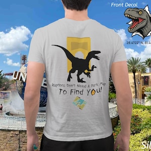 Puede incluir: Camiseta plateada con un gráfico de dinosaurio negro y el texto "Raptors Don't Need A Park Map To Find You!". Un gráfico de cabeza de dinosaurio está en la parte superior derecha. La camiseta es un recuerdo de un parque temático.