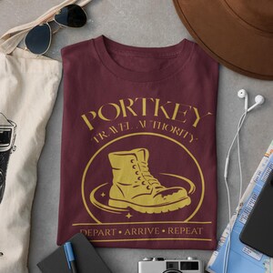 Può includere: Maglietta bordeaux con grafica dorata che recita "PORTKEY TRAVEL AUTHORITY" attorno all'illustrazione di uno stivale. La maglietta riporta anche le parole "DEPART • ARRIVE • REPEAT". Accessori: occhiali da sole, cappello e macchina fotografica.