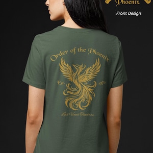 Könnte beinhalten: Ein militärgrünes T-Shirt mit einer goldenen Grafik auf der Rückseite, die einen Phönix sowie den Text Order of the Phoenix, Est. 1970 und Lux Vincit Tenebras zeigt. Oben rechts ist eine goldene Grafik mit zwei gekreuzten Zauberstäben abgebildet.