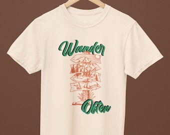 Op de afbeelding: Ivoor kleurig T-shirt met de tekst "Wander Often" in groen en oranje. Het ontwerp toont een wegwijzer met illustraties van bergen, bomen en een meer. Het T-shirt hangt aan een houten hanger.
