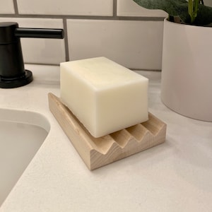 May include: A white bar of soap sits on a wooden soap dish with a wavy design. The dish is on a white countertop.