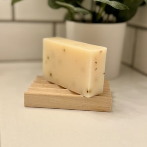 May include: A white bar of soap with brown flecks sits on a wooden soap dish. The soap dish is made of light wood and has a rectangular shape.