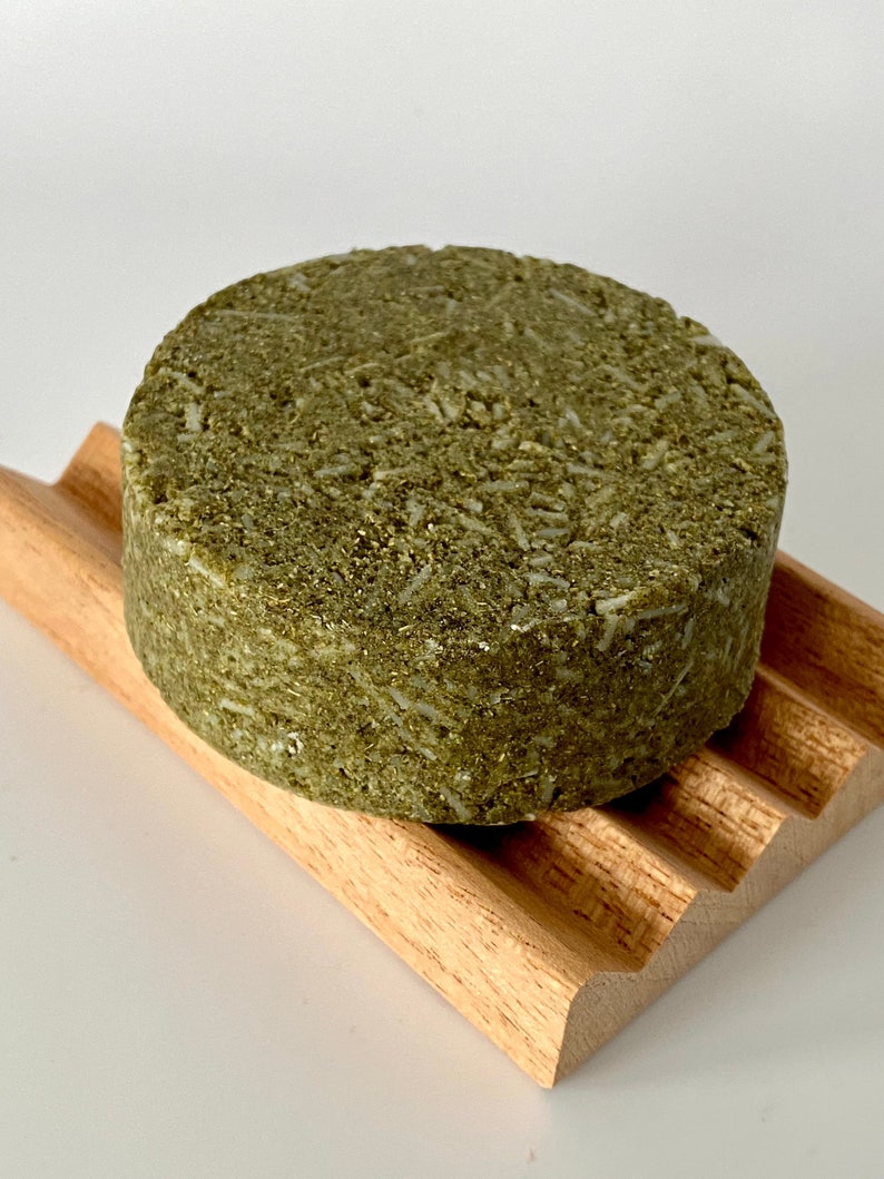 May include: A round, green, and brown shampoo bar on a wooden soap dish. The bar is made with natural ingredients and is designed to be gentle on hair and scalp.