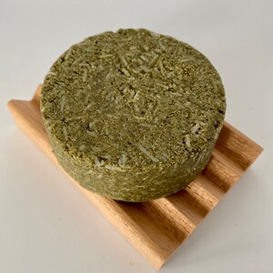 May include: A round, green and white bar of soap on a wooden soap dish. The soap is made with natural ingredients and is designed to cleanse and nourish the skin.