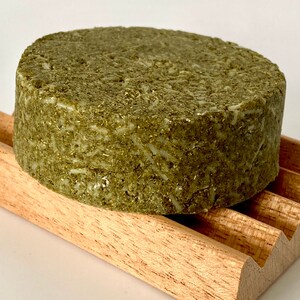 May include: A round, green, solid bar of shampoo on a wooden soap dish. The shampoo bar is made with natural ingredients and is designed to be gentle on hair and scalp.