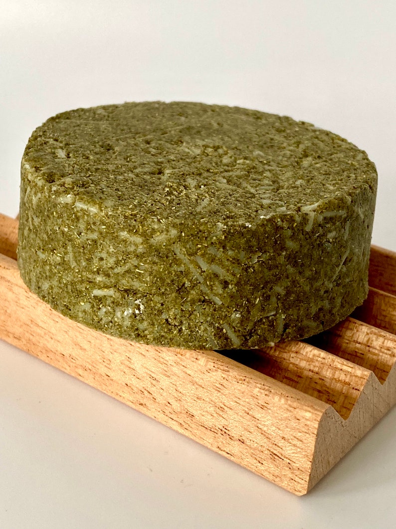 May include: A round, green, solid bar of shampoo on a wooden soap dish. The shampoo bar is made with natural ingredients and is designed to be gentle on hair and scalp.