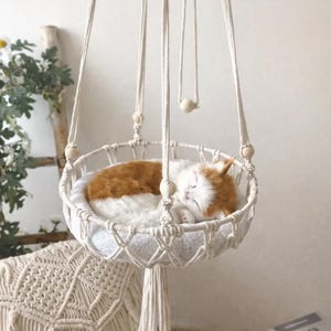 May include: A hanging macramé cat bed with a sleeping ginger and white cat. The bed is suspended by woven cords and features a round, woven base. Wooden beads accent the design, creating a cosy and stylish pet accessory.
