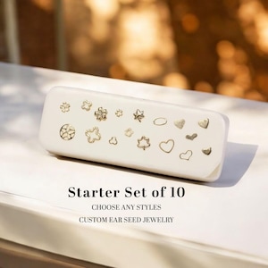 May include: A white rectangular case with various gold-colored stud earrings in different shapes, including stars, hearts, and flowers. The text "Starter Set of 10" is displayed below the case, along with "Choose Any Styles" and "Custom Ear Seed Jewelry."