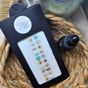 May include: A black earring display card with a variety of stud earrings. The earrings feature designs such as stars, crosses, pearls, and shells. A black bottle with a dropper is next to the display card. The card has a white label with the text "Teeny Tiny Jewelry".
