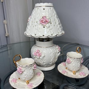 Könnte beinhalten: Eine weiße Keramiklampe mit floralem Design und zwei passenden Tassen und Untertassen. Die Lampe hat einen durchbrochenen Schirm und ist mit rosa Rosen verziert. Die Tassen und Untertassen weisen ebenfalls rosa Rosen und goldene Akzente auf.