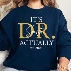 May include: Navy blue sweatshirt with the text "IT'S DR. Smith ACTUALLY est. 2004" in gold and white lettering. The sweatshirt is a crew neck style and is being worn by someone.