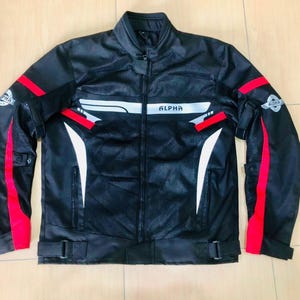 May include: Black motorcycle jacket with red and white detailing. The jacket has the word "ALPHA" across the chest and white and red stripes on the sleeves. It features a stand-up collar and a zip closure.