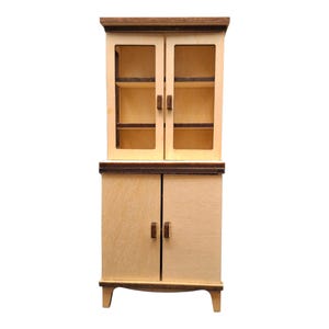 Könnte beinhalten: Ein Miniatur-Holzschrank mit hellem Holzfinish und dunkelbraunen Zierleisten. Der obere Teil hat Glastüren, während der untere Teil massive Türen hat. Der Schrank ist im klassischen Stil gehalten und eignet sich für ein Puppenhaus oder eine Miniatursammlung.