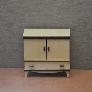 Könnte beinhalten: Ein Miniatur-Holzschrank mit zwei Türen, einer Schublade und einer abgeschrägten Oberseite. Der Schrank ist hellfarbig mit dunkelbraunen Zierleisten und Griffen. Er steht auf vier gebogenen Beinen vor einem strukturierten grauen Hintergrund.