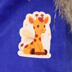 Op de afbeelding: Een sticker met een cartoon giraffe met vleugels, oranje vlekken en een rode manen. De giraffe zit met gekruiste benen. De sticker heeft een witte rand en staat op een blauwe achtergrond.