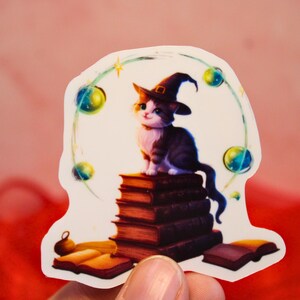 Può includere: Un adesivo raffigurante un gatto dei cartoni animati con un cappello da strega, seduto su una pila di libri. Il gatto è bianco e nero, con un'espressione allegra. L'adesivo ha un bordo bianco con un design celeste.
