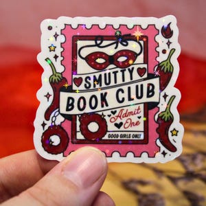 Könnte beinhalten: Ein rosa Aufkleber mit einem Stempeldesign mit dem Text "Smutty Book Club" und anderem Text. Der Aufkleber enthält eine rote Maske, Chilischoten und ein Herz. Der Aufkleber wird von einer Hand gehalten.