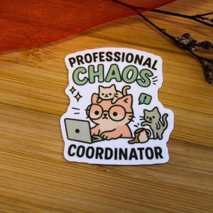 Puede incluir: Una pegatina blanca con las palabras "PROFESSIONAL CHAOS COORDINATOR". Presenta un gato de dibujos animados con gafas y un port&aacute;til, con otros dos gatos. La pegatina est&aacute; sobre una superficie de madera.