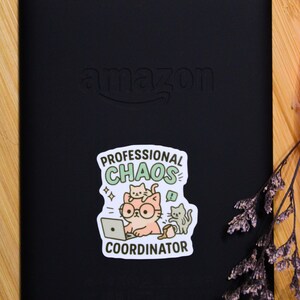 Puede incluir: Lector electr&oacute;nico negro con el logotipo de Amazon y una pegatina que dice "Professional Chaos Coordinator". La pegatina presenta un gato de dibujos animados con gafas y usando una computadora port&aacute;til, con otros dos gatos. El fondo es una superficie de madera.