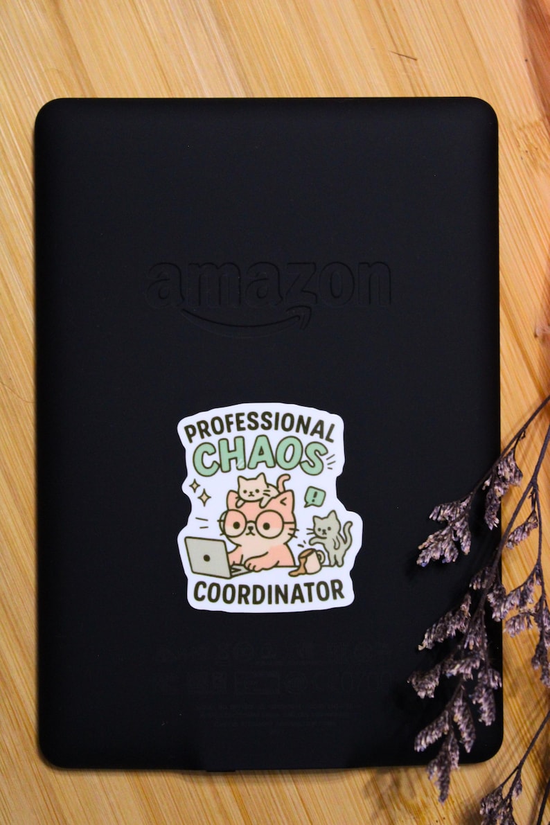 Puede incluir: Lector electr&oacute;nico negro con el logotipo de Amazon y una pegatina que dice "Professional Chaos Coordinator". La pegatina presenta un gato de dibujos animados con gafas y usando una computadora port&aacute;til, con otros dos gatos. El fondo es una superficie de madera.