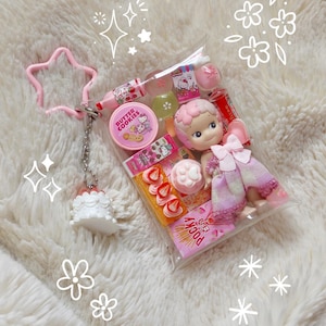 K&ouml;nnte beinhalten: Eine transparente Plastikt&uuml;te gef&uuml;llt mit Miniatur-S&uuml;&szlig;igkeiten in Rosa und Wei&szlig;, einer Puppe und Lipgloss. Die Tasche ist an einem rosa sternf&ouml;rmigen Schl&uuml;sselanh&auml;nger und einem kleinen wei&szlig;en Kuchenanh&auml;nger befestigt. Die Artikel sind auf einer flauschigen wei&szlig;en Oberfl&auml;che angeordnet.