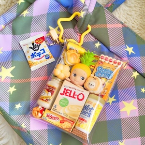 May include: A clear plastic bag filled with miniature food items, including a Jell-O box, a bottle of Alfredo sauce, and a snack bag with Japanese text. The bag is attached to a yellow star-shaped keychain, set against a plaid tote bag.
