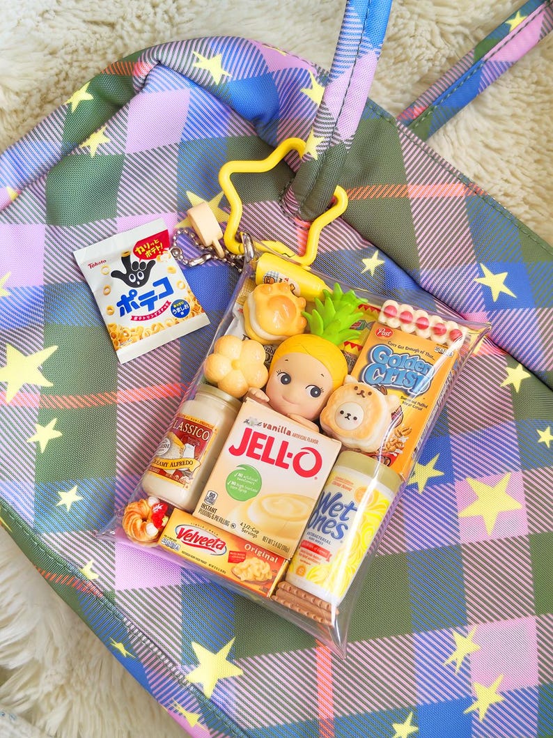 May include: A clear plastic bag filled with miniature food items, including a Jell-O box, a bottle of Alfredo sauce, and a snack bag with Japanese text. The bag is attached to a yellow star-shaped keychain, set against a plaid tote bag.