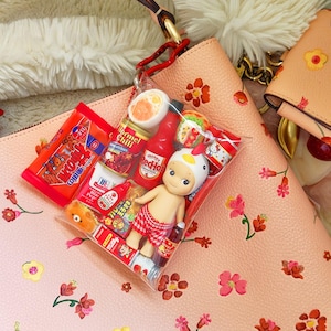 May include: A clear plastic bag filled with miniature food items and a small doll wearing a chicken hat and red checkered shorts. The bag is hanging from a peach-colored purse with floral designs. Other peach-colored purses are in the background.