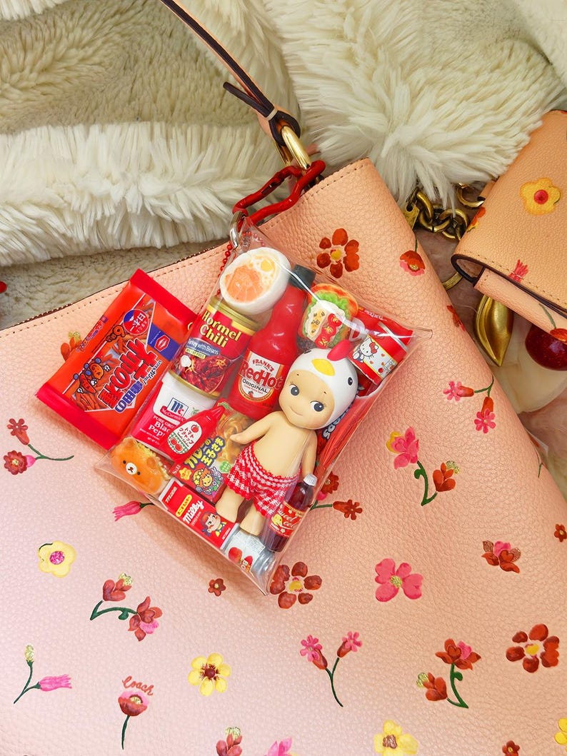May include: A clear plastic bag filled with miniature food items and a small doll wearing a chicken hat and red checkered shorts. The bag is hanging from a peach-colored purse with floral designs. Other peach-colored purses are in the background.