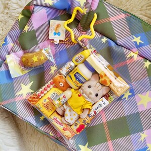May include: A plaid tote bag in shades of green, pink, and blue, adorned with yellow stars. A clear plastic bag contains miniature food items and plush toys. A yellow star-shaped keychain and a small tag are attached to the bag.