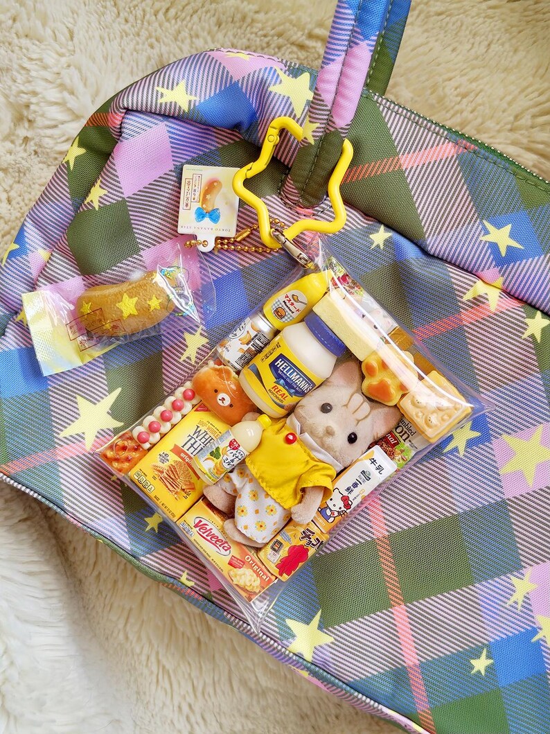 May include: A plaid tote bag in shades of green, pink, and blue, adorned with yellow stars. A clear plastic bag contains miniature food items and plush toys. A yellow star-shaped keychain and a small tag are attached to the bag.