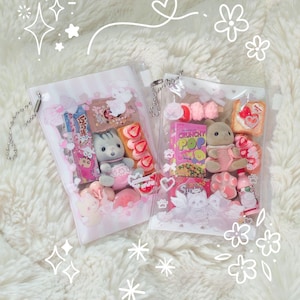 K&ouml;nnte beinhalten: Zwei transparente Beutel gef&uuml;llt mit verschiedenen rosa und wei&szlig;en S&uuml;&szlig;igkeiten, Miniatur-Pl&uuml;schtieren und Snacks. Die Beutel sind mit Blumen- und Herzmotiven verziert und liegen auf einer flauschigen wei&szlig;en Oberfl&auml;che. Ein Beutel hat eine silberne Kette.