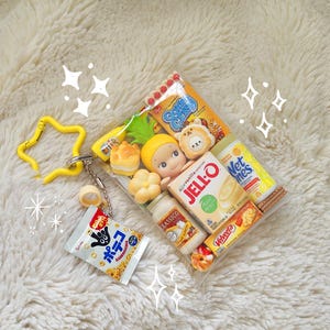 May include: A clear, rectangular pouch filled with miniature food items and figurines. Includes a small bag of popcorn, a bottle of yogurt, a box of Jell-O, and other snacks. A yellow star-shaped keychain is attached to the pouch.