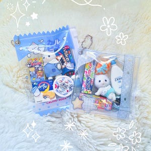 May include: Two clear, blue-trimmed pouches filled with assorted items. One contains a plush cat, snacks, and a drink. The other has a plush bear, snacks, and a drink. The pouches are on a fluffy white surface.