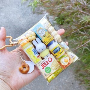 Peut inclure: Une pochette rectangulaire transparente remplie de figurines miniatures de nourriture et de personnages, dont Jell-O, de la mayonnaise Hellmann's, Crystal Light et un lapin Miffy. Un petit beignet marron est suspendu &agrave; une cha&icirc;nette argent&eacute;e.