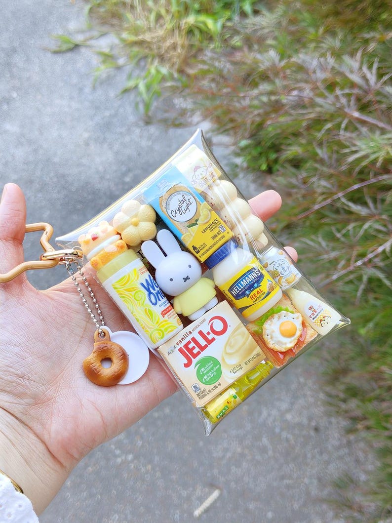Peut inclure: Une pochette rectangulaire transparente remplie de figurines miniatures de nourriture et de personnages, dont Jell-O, de la mayonnaise Hellmann's, Crystal Light et un lapin Miffy. Un petit beignet marron est suspendu &agrave; une cha&icirc;nette argent&eacute;e.