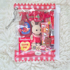 Puede incluir: Una bolsa de pl&aacute;stico transparente con un borde a cuadros rojo y blanco, llena de varios objetos peque&ntilde;os. Incluye un conejito de peluche, caramelos, una peque&ntilde;a botella de jarabe y otros juguetes en miniatura. La bolsa tiene una cadena plateada.