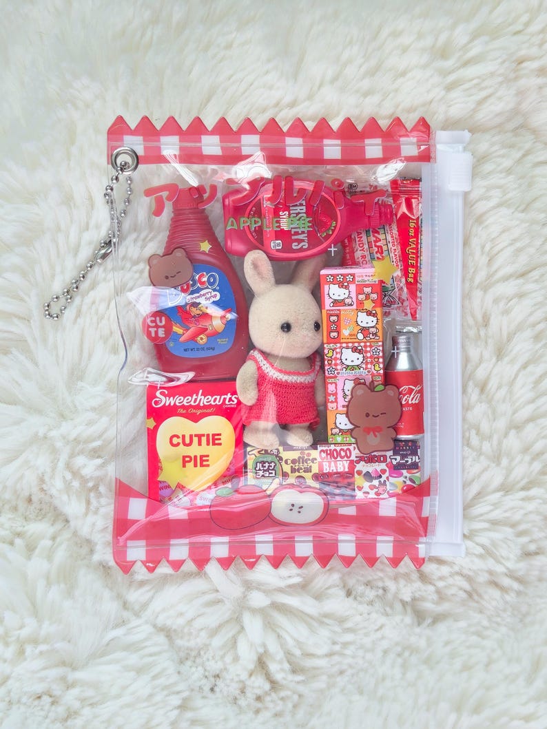Puede incluir: Una bolsa de pl&aacute;stico transparente con un borde a cuadros rojo y blanco, llena de varios objetos peque&ntilde;os. Incluye un conejito de peluche, caramelos, una peque&ntilde;a botella de jarabe y otros juguetes en miniatura. La bolsa tiene una cadena plateada.