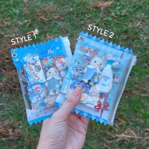 以下が含まれることがあります： 「STYLE 1」と「STYLE 2」とラベルされた、青いトリムが付いた2つの透明なビニール袋。ミニチュアのおもちゃとスナックが詰められています。緑の草を背景に袋が持たれています。袋にはミニチュアのおもちゃ、お菓子、Doveのボトルが入っています。