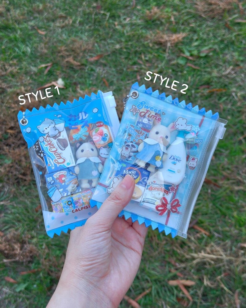 以下が含まれることがあります： 「STYLE 1」と「STYLE 2」とラベルされた、青いトリムが付いた2つの透明なビニール袋。ミニチュアのおもちゃとスナックが詰められています。緑の草を背景に袋が持たれています。袋にはミニチュアのおもちゃ、お菓子、Doveのボトルが入っています。