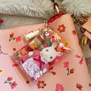 Puede incluir: Un bolso de mano de cuero rosa con un estampado floral, que contiene un charm de bolsa de pl&aacute;stico transparente. El charm contiene un conejito de peluche, alimentos en miniatura y una lata peque&ntilde;a. El bolso tiene un asa roja y herrajes dorados.