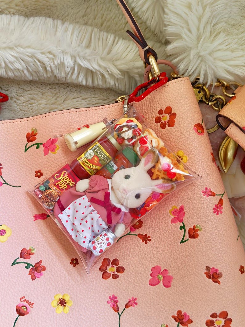 Puede incluir: Un bolso de mano de cuero rosa con un estampado floral, que contiene un charm de bolsa de pl&aacute;stico transparente. El charm contiene un conejito de peluche, alimentos en miniatura y una lata peque&ntilde;a. El bolso tiene un asa roja y herrajes dorados.