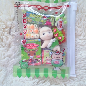 May include: A clear plastic bag filled with Japanese snacks and a small plush toy. The bag contains colorful candies, a drink illustration, and a small plush animal. The bag has a green zig-zag trim and a silver chain.