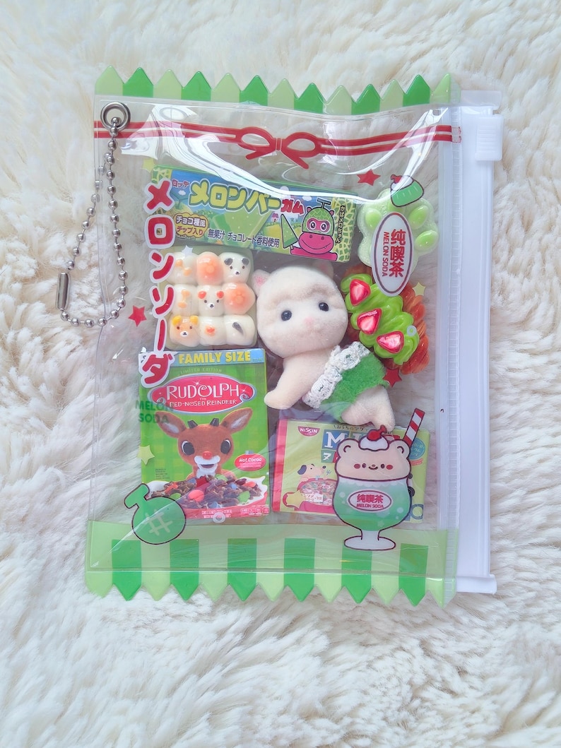 May include: A clear plastic bag filled with Japanese snacks and a small plush toy. The bag contains colorful candies, a drink illustration, and a small plush animal. The bag has a green zig-zag trim and a silver chain.