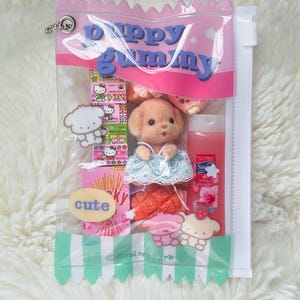 May include: A clear plastic bag with a pink top and a green and white striped bottom, filled with various items. The bag contains a puppy doll, candies, and a bottle of liquid. The text "Puppy Gummy" is printed on the bag.