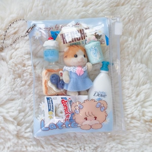 May include: A clear plastic pouch filled with miniature toys and accessories. The pouch contains a doll in a blue dress, a bottle of baby Dove, a Hershey's cookie, a cupcake, and other small items. The text "always by your side" is printed on the pouch.