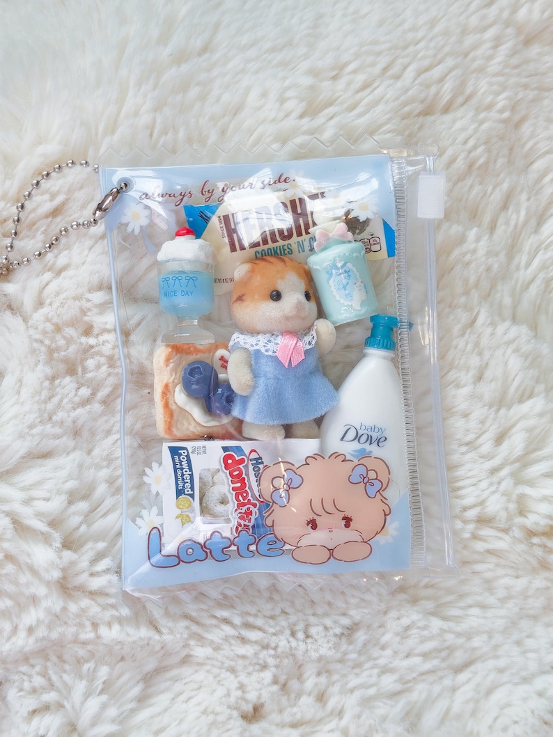 May include: A clear plastic pouch filled with miniature toys and accessories. The pouch contains a doll in a blue dress, a bottle of baby Dove, a Hershey's cookie, a cupcake, and other small items. The text "always by your side" is printed on the pouch.